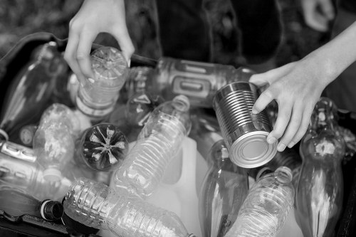 A black-and-white photograph showing a personâ€™s hands sorting recyclable waste outdoors. One hand is gripping a small, crushed, metal food can with a smooth, cylindrical shape and a reflective surface, while the other hand is holding a plastic bottle with ridges along its body and a screw-top lid. Surrounding the hands are various discarded items, including multiple plastic bottles of different sizes and shapes, some with labels and caps, and several plastic containers, all lying on a flat surface. The scene suggests waste sorting or collection, with the focus on identifying recyclable materials such as plastics and metals. The background appears to show an outdoor environment with natural light, possibly a garden or a collection point for rubbish disposal, aligning with the services provided by Office Clearance Poplar for rubbish removal near Poplar, London, which operates in the E14 postcode. The overall texture highlights the different materials, including the shiny surfaces of the cans and bottles and the matte finish of certain containers.
