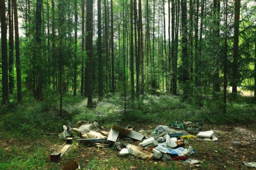 In a woodland area with tall, densely packed trees featuring green foliage, a pile of discarded household waste is situated on the forest floor. The rubbish includes various materials such as cardboard boxes, plastic bags, and other miscellaneous debris, some of which are crushed or torn, reflecting improper disposal in a natural environment. The waste appears to be scattered and haphazardly stacked on a patch of bare soil amidst the trees, with minimal ground vegetation visible around it. The lighting indicates daylight filtering through the leafy canopy, illuminating the scene with soft natural light. This image highlights the issue of illegal dumping and environmental pollution, consistent with the kind of rubbish removal services offered by companies like Office Clearance Poplar in the Poplar area. Such scenes are encountered when waste is not disposed of properly, emphasizing the importance of professional clearance services to maintain clean and safe surroundings.