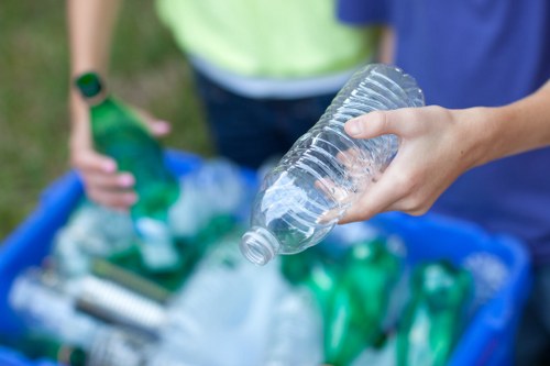 A close-up of a person's hand holding an empty, clear plastic bottle with a faint ribbed texture, positioned in the foreground. In the background, another individual wearing a high-visibility yellow vest and a wristwatch is seen holding a green plastic bottle, standing outdoors. A blue plastic container filled with various discarded plastic bottles and containers, including green and transparent ones, rests on a paved surface, likely part of a rubbish collection or disposal process near a location such as Poplar in London. The scene highlights waste materials primarily composed of plastic, consistent with rubbish removal services offered by companies like Office Clearance Poplar. Natural daylight illuminates the scene, emphasizing the different textures and colors of the waste items and the setting's outdoor environment. The overall image conveys the process of sorting or preparing plastic waste for collection or recycling.
