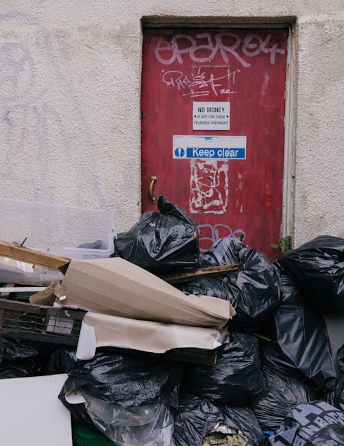 A large pile of metal debris and construction waste situated in front of an unfinished or abandoned building with multiple large windows. The debris includes twisted metal rods, broken panels, and dis