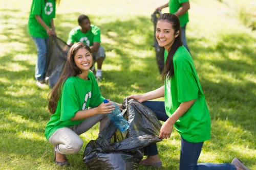Two young women wearing bright green recycling T-shirts are kneeling on a well-maintained grassy area outdoors, actively participating in a community cleanup event. The woman on the left, with long brown hair, is smiling while holding a large black plastic rubbish bag open, ready to deposit waste. She has beige trousers and the T-shirt displays a white recycling symbol. The woman on the right, also smiling, has long dark hair and is wearing dark jeans; she is pulling the bag toward her. In the background, three other volunteers, also dressed in green T-shirts, are engaged in collecting more litter and holding additional black rubbish bags. They are positioned on a sunny day with dappled shade from trees, in an open, park-like setting, suggesting a positive community effort to promote environmental responsibility. This scene reflects a waste management initiative typical of local eco-friendly activities, with a focus on recycling and clean-up work, relevant to community and environmental services such as rubbish removal in the Poplar area.